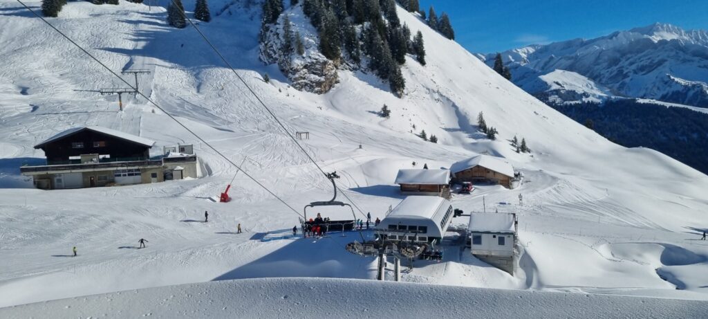 Im Vordergrund eine Schneekante, darüber im Sessellift sitzt jemand mit zwei Kindern, im Hintergrund beginnt die Abfahrt auf der Piste entlang der Zahnradbahn und endet eine kurze Strecke entlang des Bergs Roc a l'Ours.