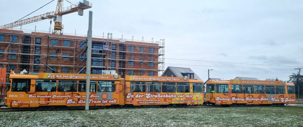 Von der Seite und in voller Breite, Gelenkwagen 7756 und Beiwagen 7772 im orangenen 1970er Flower Power-Design und mit lustigen Sprüchen