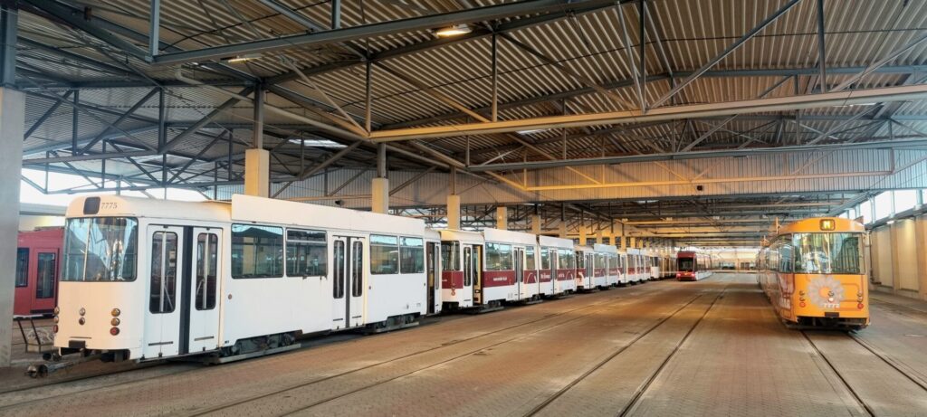 Eine breite Halle mit Dachkonstruktion und Wellblechdach, links der weiße Duewag-Triebwagen 7775 vor weiteren mit dunkelroten Streifen, rechts der orangene Duewag-Beiwagen 7772 im 1970er Flower Power-Design