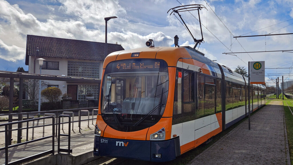 Dunkelblaue Schürze mit rnv-Logo und Wagennummer 5711 unter orangenem Aufbau rund um die Frontscheibe, im Weiterem weitgehend Weiß mit orangenen und dunkelblauen Akzenten. Rechts daneben das Haltestellenschild "Käfertaler Wald Steig B", links im Hintergrund eine Straßenlaterne und ein zweistöckiges Haus mit Glasbausteinen am Treppenhaus