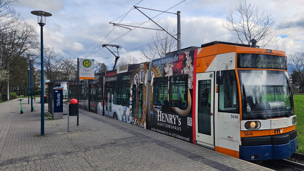 Dunkelblaue Schürze, darüber ein orangener Streifen mit rnv-Logo und Wagennummer 5646, darüber ein weißer Streifen mit den Scheinwerfern, darüber orange umfasst die Frontscheibe und die digitale Zielanzeige, die restliche Straßenbahn beklebt mit Werbung für ein Aktionshaus. Links daneben das Haltestellenschild "Vogelstang Steig A", ein Mülleimer, ein Fahrkartenautomat und ein Wartehäuschen, außerdem Laternen und zwei E-Scooter.