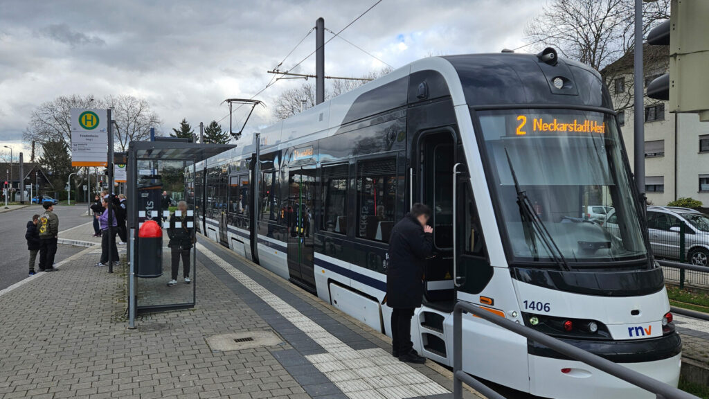 Nicht so breit aber höher als die alten Straßenbahntypen, weiß mit schwarzen Elementen und tief nach unten gezogener, schrägen Frontscheibe. Die Fahrerin raucht in ihrer Pause, links neben der Straßenbahn ein Mülleimer, ein Wetterschutz, ein Haltestellenschild für die Busse am Steig links daneben, einige wartende Menschen.