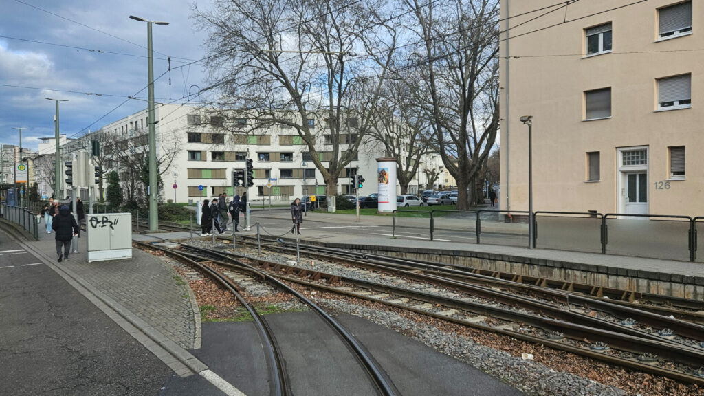 Zwei Gleise zweigen ab, im Hintergrund links der regulär von den Linien 6 und 6A bediente Bahnsteig in Fahrtrichtung Rheingönheim, davor überqueren Menschen die Gleise an einem Ampel-gesicherten Übergang