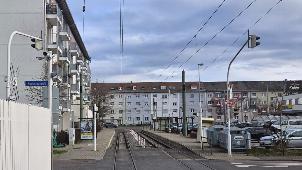 Links vorne im Bild ein weiß gestrichener Stahlzaun, dahinter kreuzt die Sudermannstraße Ampel-gesichert die Betriebsstrecke, dahinter links und rechts zwei Bahnsteige inklusive Werbung. Am Bahnsteig links E-Scooter, am Bahnsteig rechts Glas- und Kleidercontainer. Im Hintergrund das Gleisdreieck und die Mehrfamilienhäuser der Saarlandstraße.