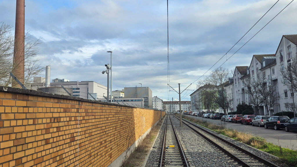 Links eine Mauer aus hellbraunen Steinen mit Stacheldraht und Videoüberwachung, in der Mitte die beiden Gleise, rechts die Richard-Dehmel-Straße mit geparkten Autos