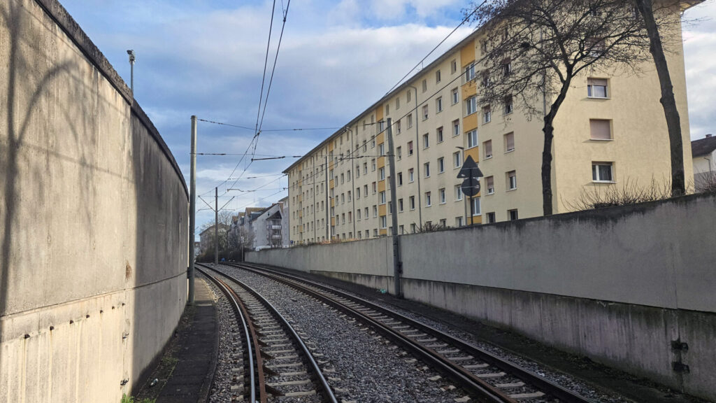 Die Schienen beschreiben eine leichte Kurve nach links, links von ihnen eine höhere Mauer zum Abbvie-Werk als rechts zur Straße. Neben der Straße zunächst ältere, dann neuere Mehrfamilienhäuser.