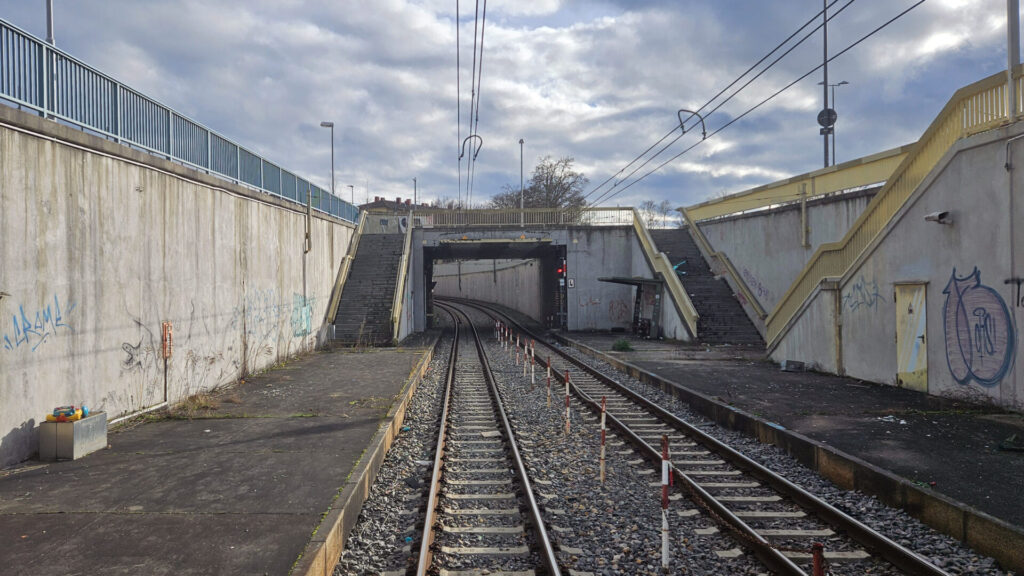 Alles sehr herunter gekommen: viel beschmierter Beton an den Wänden links und rechts, flache geteerte Bahnsteige, im Hintergrund führen Treppen hoch zu einer Straßenbrücke, auf der rechten liegt ein E-Scooter. Rot-weiße, mit einer Kette verbundene Pfosten sollen das Überqueren der Gleise verhindern.