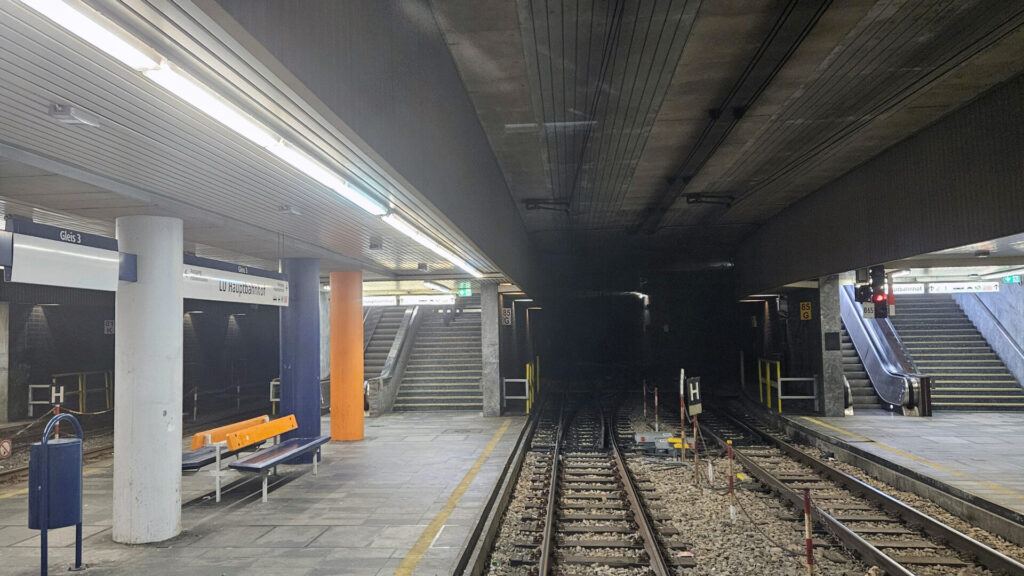 Die typische deutsche unterirdische Haltestelle mit verkleideten, runden Stützsäulen, Leuchtstoffröhren zur Beleuchtung, dunklen Wänden. Flache Bahnsteige, ein Mittelbahnsteig links, ein Außenbahnsteig rechts.