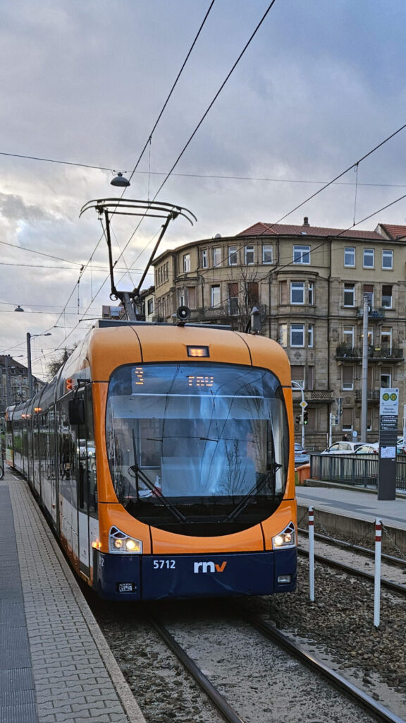 Dunkelblaue Schürze mit rnv-Logo und Wagennummer 5711 unter orangenem Aufbau rund um die Frontscheibe. Rechts verhindern dem Wagen trennen weiß-rote Pfosten mit einer Kette das Überqueren der Gleise, am Bahnsteig rechts steht ein Haltestellenschild der Doppelhaltestelle.